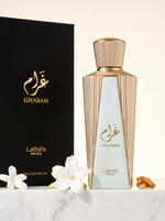 Lattafa Pride Gharam Eau de Parfum 100 ml - goldener Flakon mit Verpackung auf hellem Marmor mit weissen Blueten
