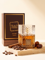 Lattafa Khamrah Qahwa Eau de Parfum 100 ml - Flakon mit Verpackung auf Sandstein mit Kaffeebohnen und Datteln