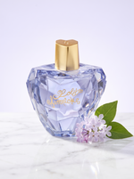 Lolita Lempicka Mon Premier Eau de Parfum 100 ml - facettierter Apfelflakon auf hellem Marmor mit Flieder