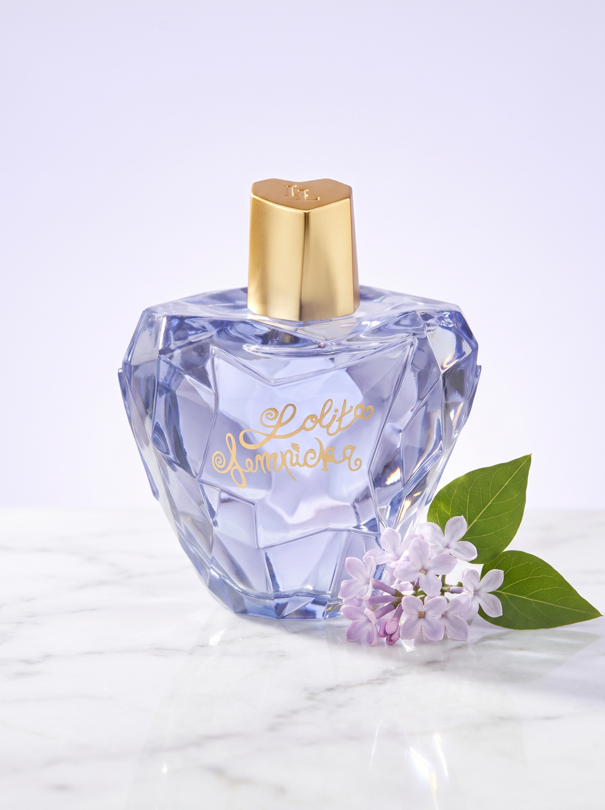 Lolita Lempicka Mon Premier Eau de Parfum 100 ml - facettierter Apfelflakon auf hellem Marmor mit Flieder