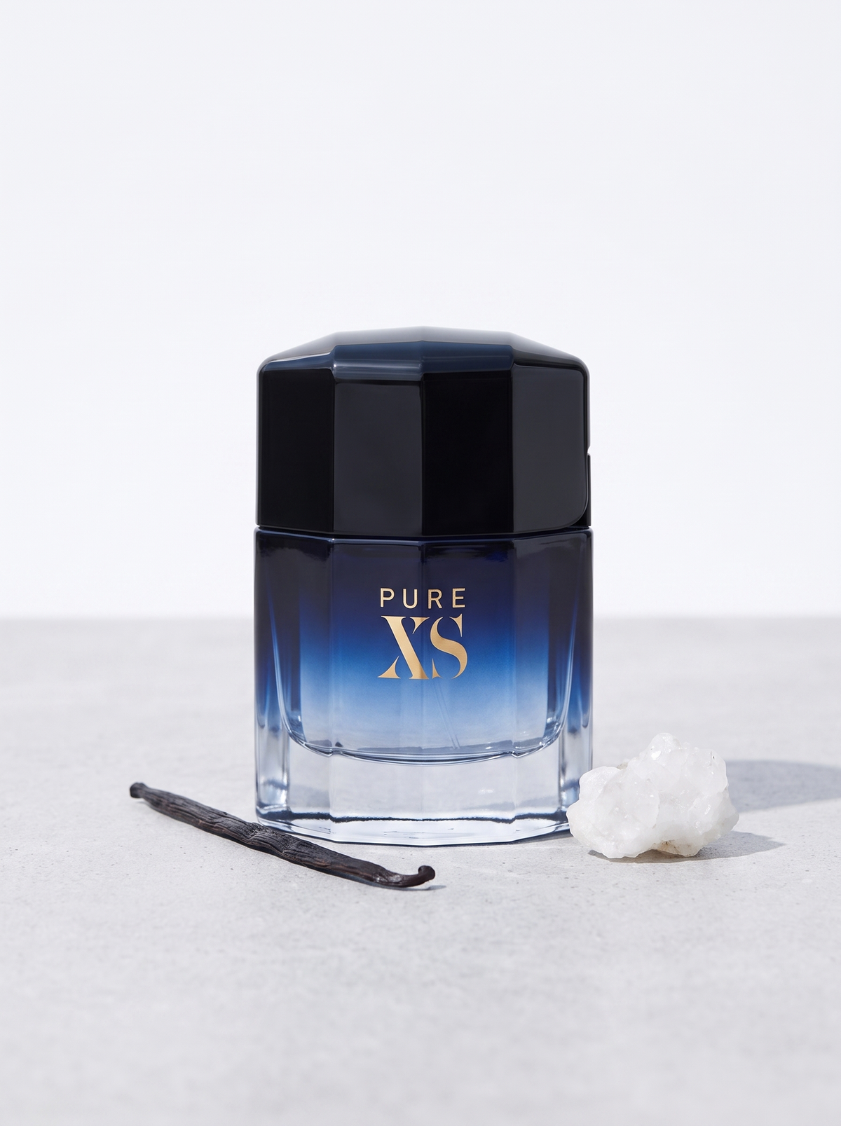 Rabanne Pure XS Eau de Toilette 100 ml - blau-schwarzer Flakon auf hellem Beton mit Vanille