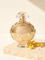Rabanne Olympea Solar Eau de Parfum Intense 80 ml - goldener Flakon auf hellem Sandstein mit Mimosen