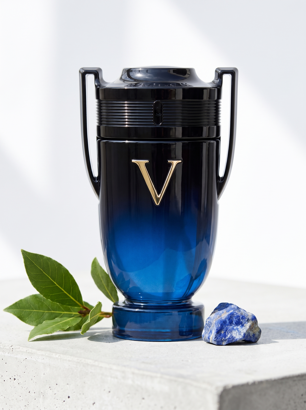 Rabanne Invictus Victory Elixir Eau de Parfum 100 ml - Pokal-Flakon auf hellem Beton mit Lorbeer und Lapislazuli